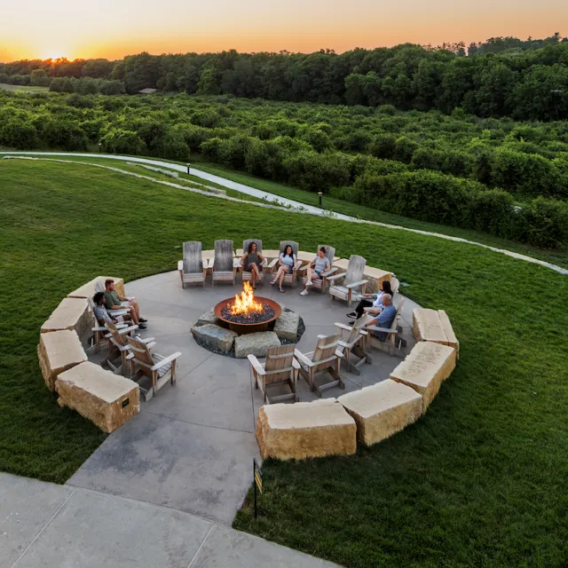 A group of people sit around a large circular fire pit on a grassy lawn with trees and a setting sun in the background.