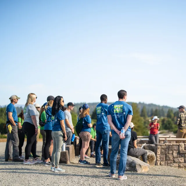 A group of people in blue shirts gather around a stone fire pit outdoors, attentively listening to a speaker. Trees and hills in the background.
