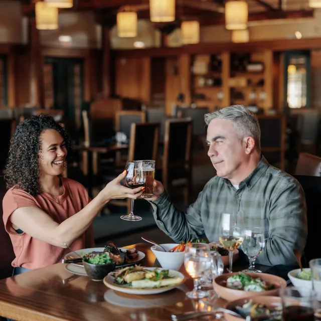 Two people toast with glasses in a cozy restaurant setting, surrounded by plates of food and soft lighting.