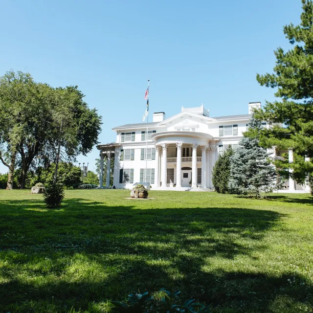 A stately white mansion with columns, surrounded by lush greenery under a clear blue sky, featuring an American flag out front.