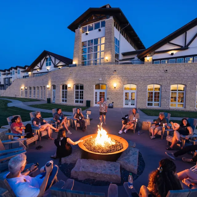 A group of people gathered around a fire pit at dusk, with a modern lodge in the background, enjoying a warm and social atmosphere.