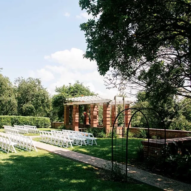 A picturesque outdoor wedding venue featuring white chairs set up on a grassy lawn, with a brick pergola and greenery in the background.
