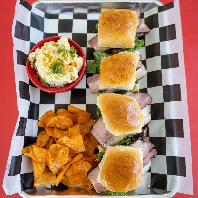 Three mini sandwiches with smoked meat on a bed of lettuce, accompanied by a bowl of creamy potato salad and a side of crispy chips.