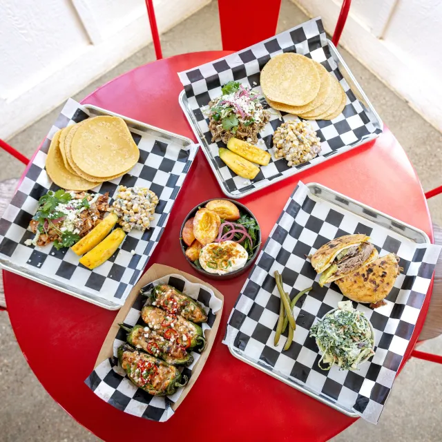A vibrant red table displays an assortment of delicious Mexican dishes, including tacos, grilled meats, jalapeños, and sides.