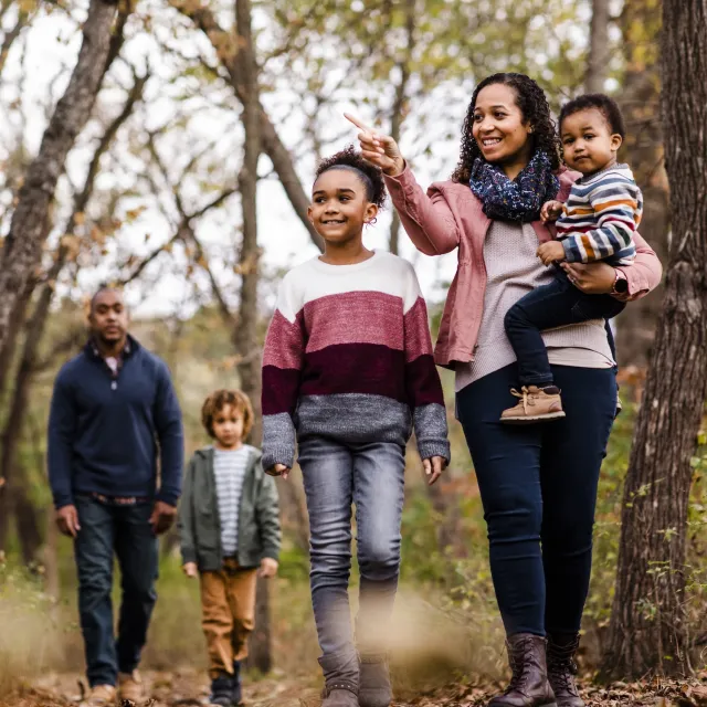 A family of five walks together on a wooded path, with trees displaying autumn foliage; one adult points out something to the children.