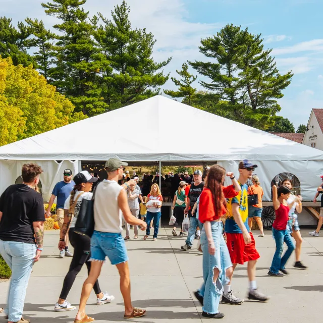 A lively outdoor event with a large white tent, surrounded by people walking in casual summer attire under a blue sky and green trees.