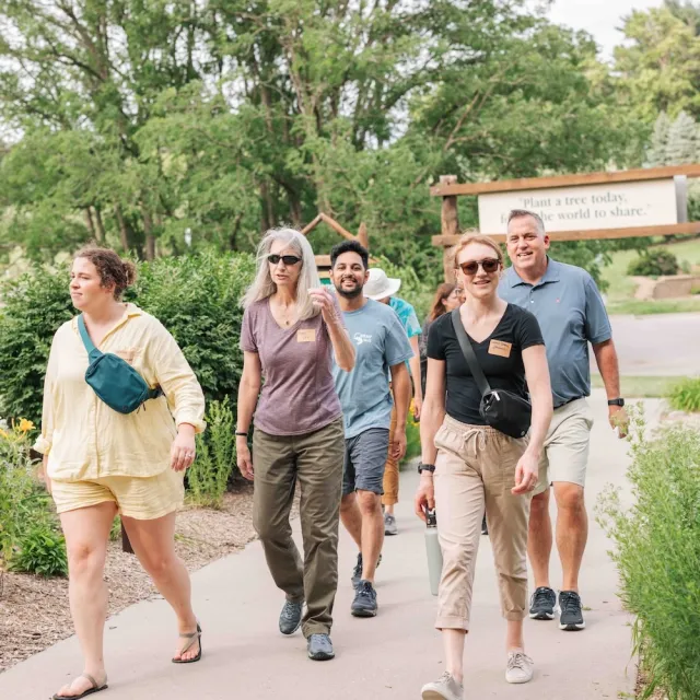 A group of five people stroll down a pathway surrounded by greenery, with a sign in the background promoting tree planting.