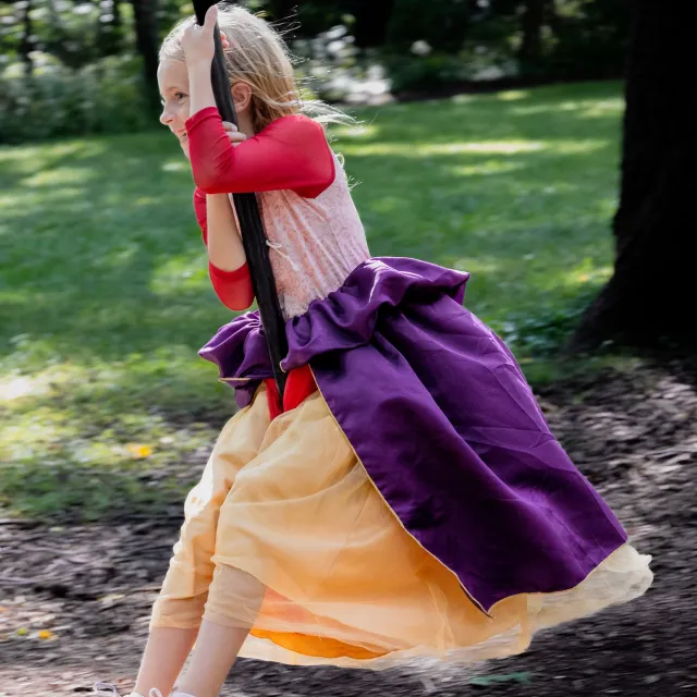 A child swings joyfully in a colorful, layered dress with purple and yellow hues, surrounded by a lush green park.