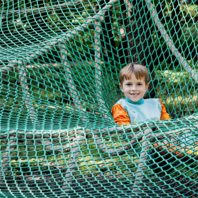 A child sits comfortably in a green net climbing structure surrounded by trees, enjoying a playful moment in nature.