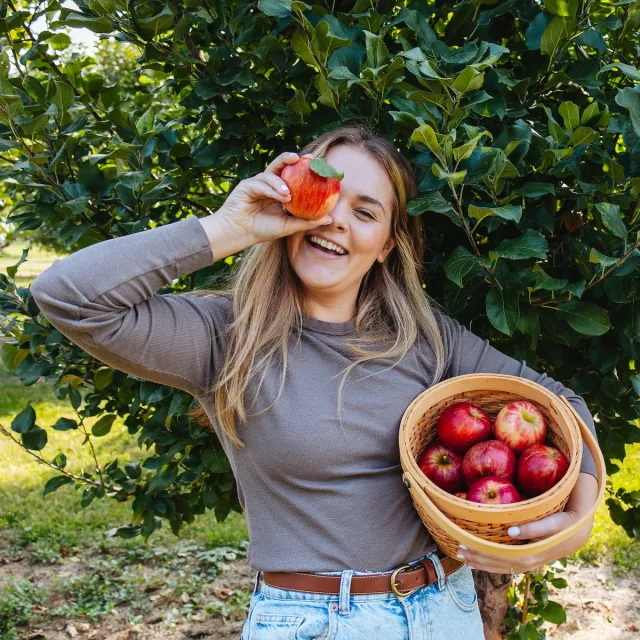 A person stands in front of an apple tree, holding a basket of freshly picked red apples against a vibrant green background.