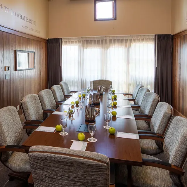 A spacious conference room featuring a long table set with glasses, apples, and papers, surrounded by comfortable chairs and bright curtains.