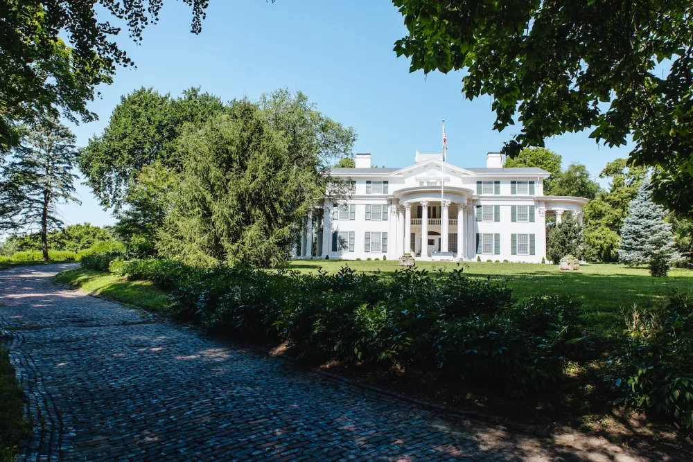 A grand white mansion with columns, surrounded by lush greenery and a paved pathway under a clear blue sky.