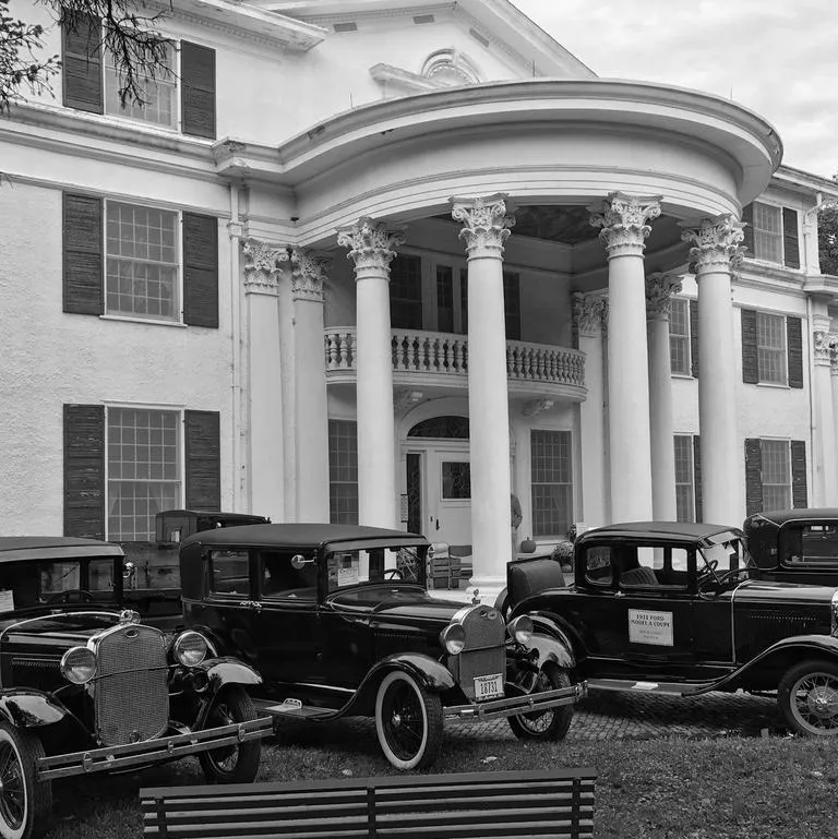 A black and white photo of vintage cars parked in front of a grand, columned mansion with large windows and an elegant entrance.