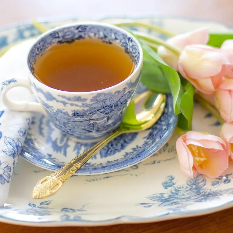 A blue and white porcelain teacup filled with tea, set on an ornate plate alongside a gold spoon and pink tulips.
