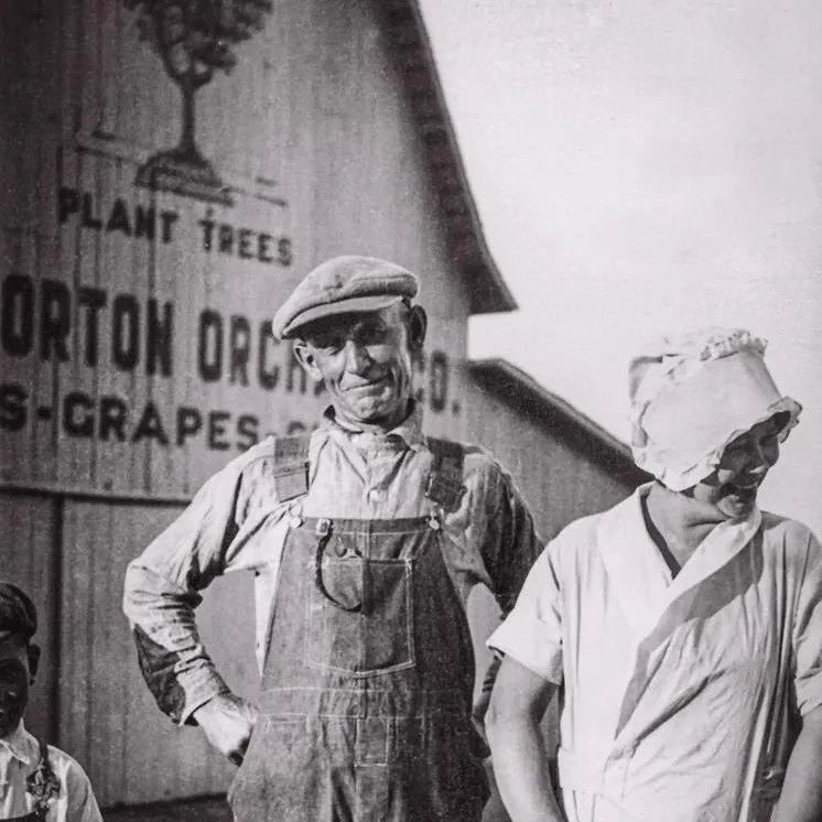 A man in overalls and a cap stands next to a woman in a white dress, with a child in front, against a barn's vintage advertisement.