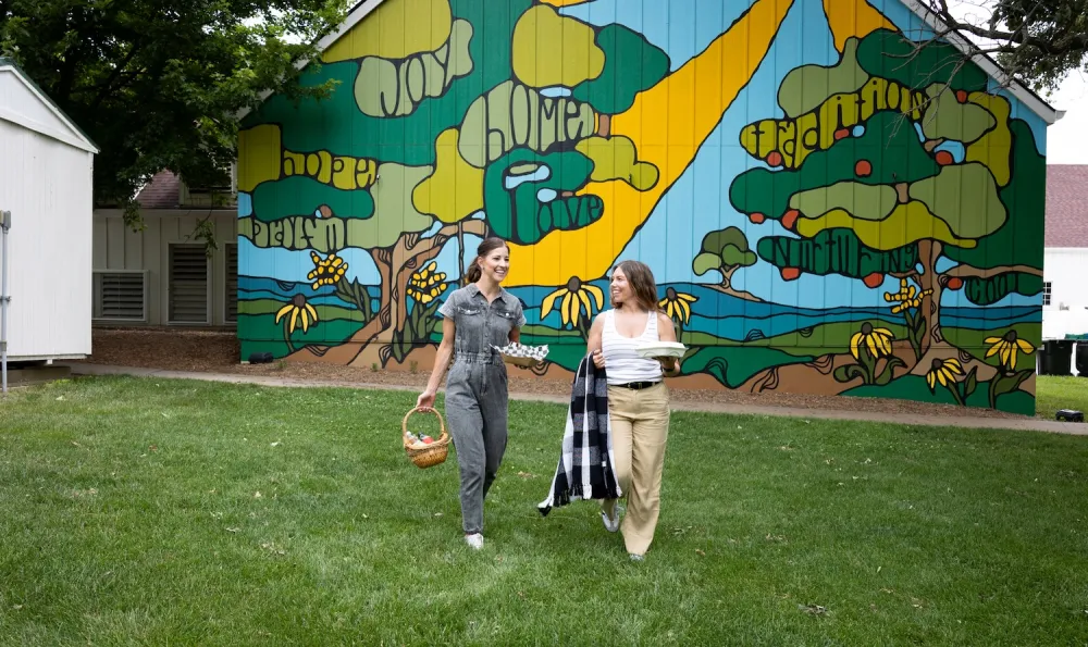 Two women walk on green grass, carrying a basket and a plate, with a colorful mural of trees and landscape behind them.