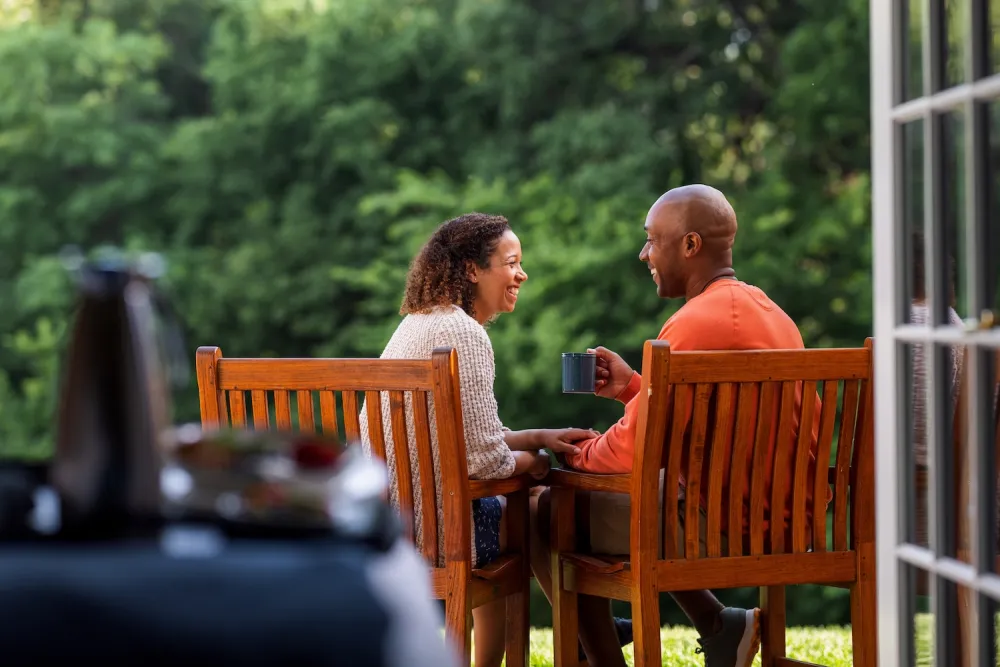 A couple sits closely on a wooden patio, enjoying coffee together, surrounded by lush greenery in a peaceful outdoor setting.