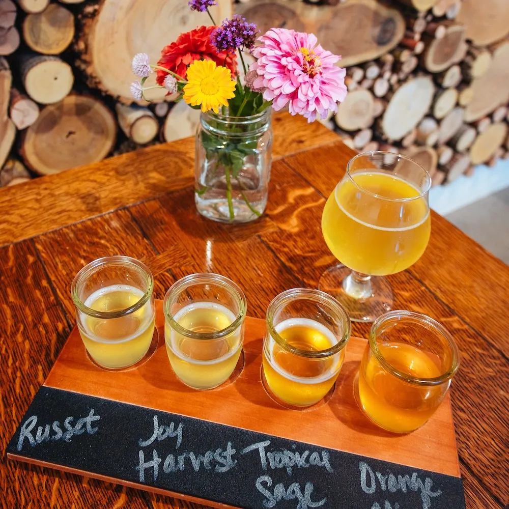 A tasting flight of four light drinks labeled 'Russet', 'Joy', 'Tropical Sage', and 'Orange' beside a floral vase on a wooden table.