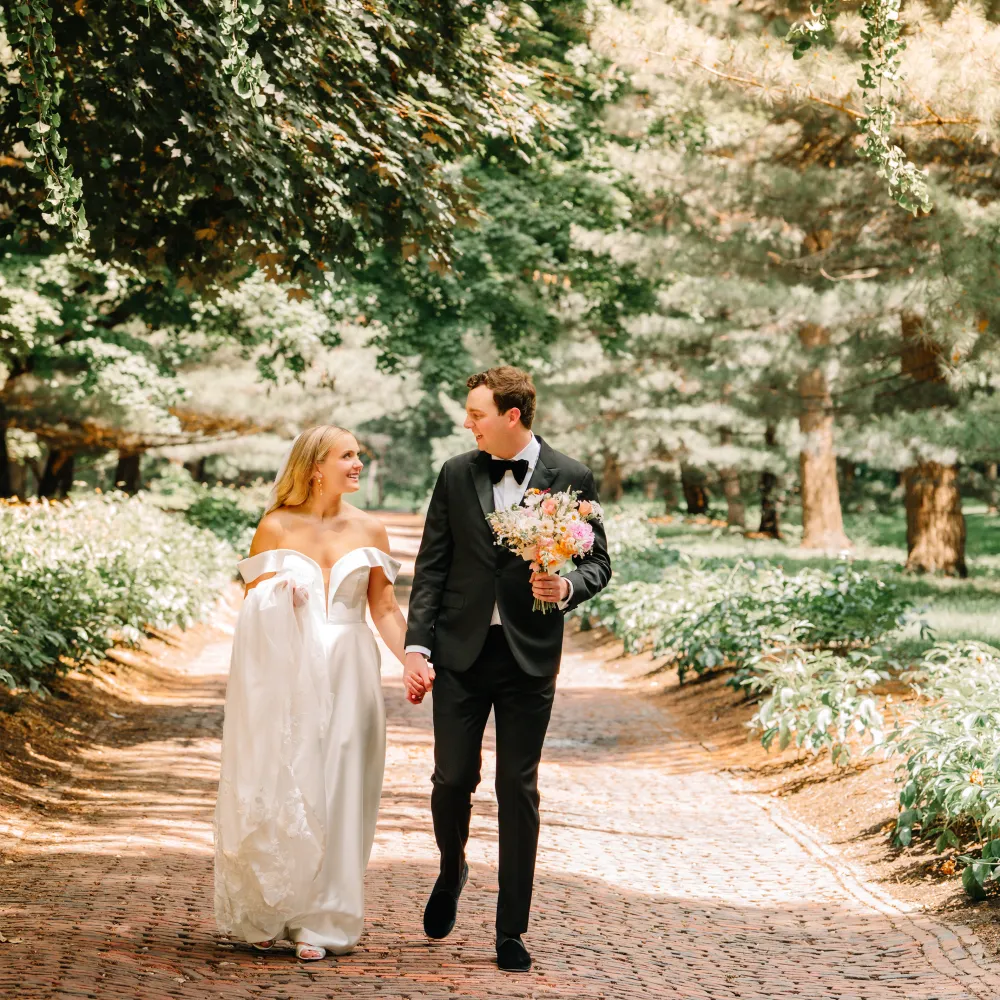 A couple walks hand in hand along a picturesque path, surrounded by greenery, the bride in a flowing white gown, the groom in formal attire.