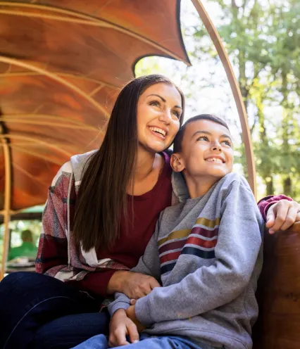 A woman and a child sit closely together on a wooden bench illuminated by soft sunlight, surrounded by lush greenery.
