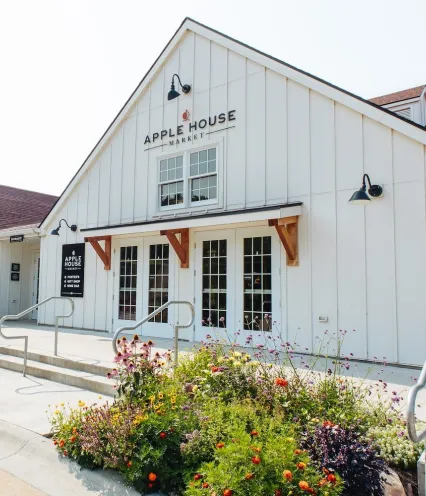 Exterior of Apple House Market featuring a large white building, colorful flower beds, and glass double doors.