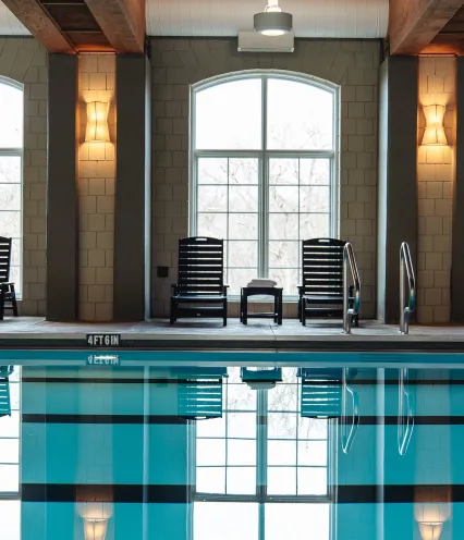 Indoor swimming pool with reflections, featuring lounge chairs, large windows, and warm lighting.