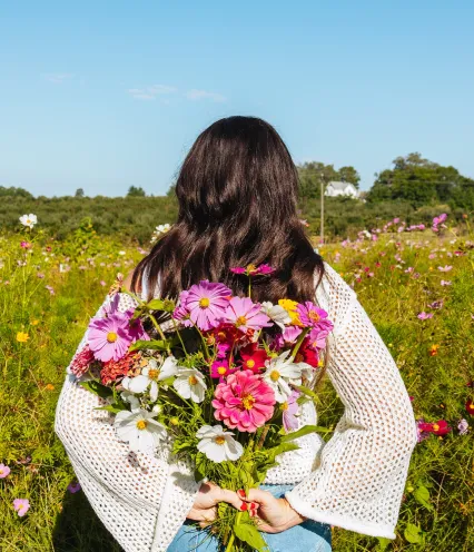 A woman in a white sweater and jeans holds vibrant pink, purple, and white flowers behind her back, standing in a colorful wildflower field under a clear blue sky.
