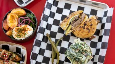 A colorful meal featuring a beef sandwich, a salad with peaches and poached egg, and stuffed jalapeños on a checkerboard tray.