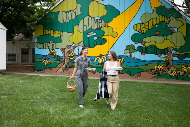 Two women walk on green grass, carrying a basket and a plate, with a colorful mural of trees and landscape behind them.