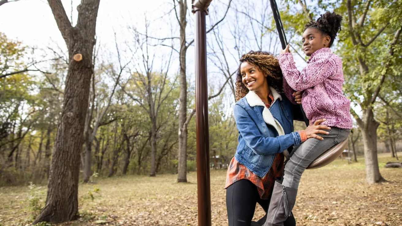 A woman and her daughter enjoying a playful moment on a swing in a vibrant park, with trees and sunlight around them.
