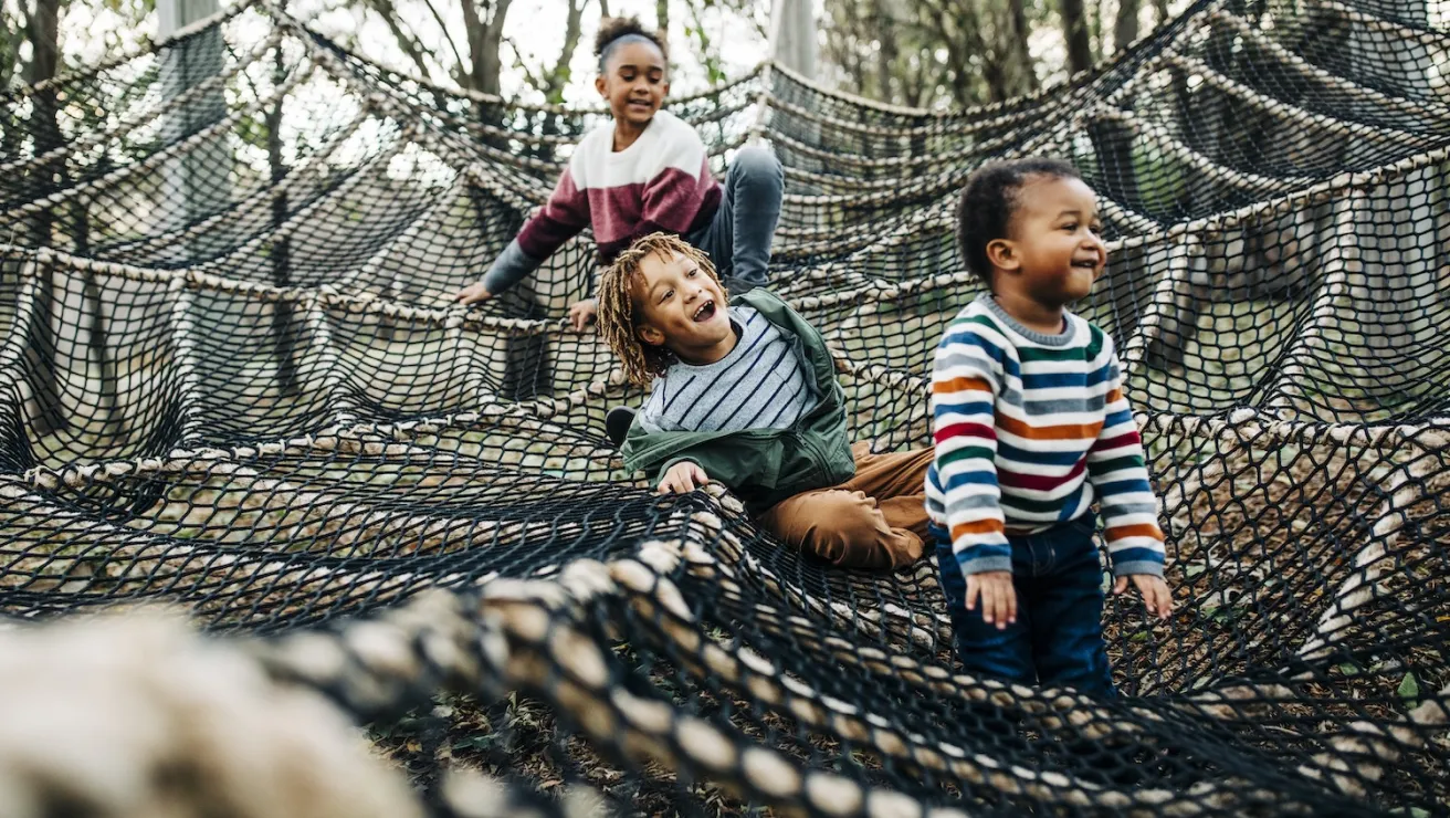 Three children play on a large outdoor net structure, surrounded by trees, engaged in a fun and adventurous activity.