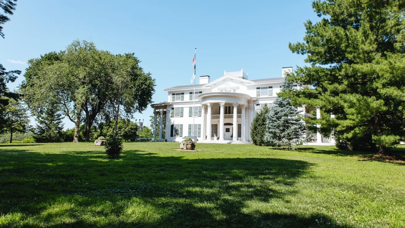 A stately white mansion with columns, surrounded by lush greenery under a clear blue sky, featuring an American flag out front.