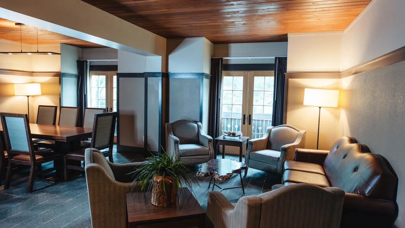 Cozy lounge area featuring two armchairs, a leather sofa, a wooden dining table, and large windows with natural light.