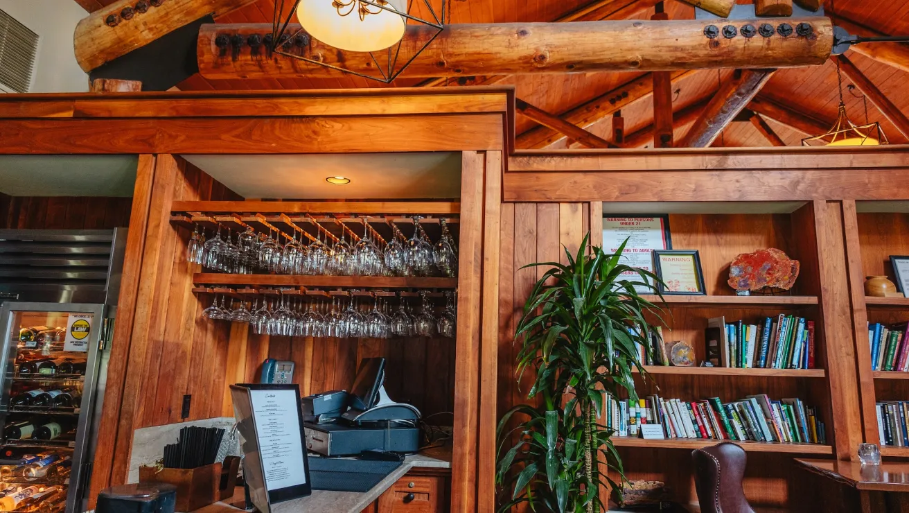 Warm wooden interior of a restaurant bar with hanging wine glasses, a cashier station, bookshelves, and a green plant. Cozy ambiance.
