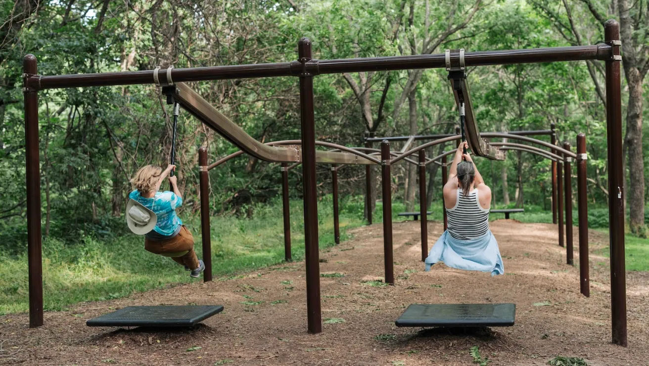 Two women swing on parallel playground bars in a lush, green forest, enjoying a sunny day of outdoor fun.