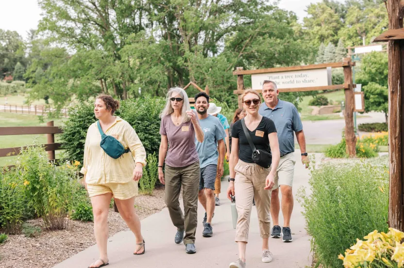 A group of five people walks along a pathway in a lush green environment, with trees and flowers surrounding them.