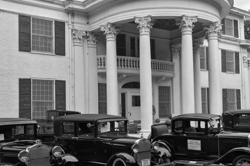 A black and white photo of vintage cars parked in front of a grand, columned mansion with large windows and an elegant entrance.