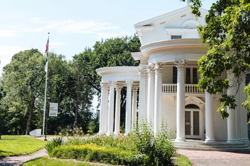 A grand white mansion with Corinthian columns, surrounded by greenery, under a blue sky, showcasing American flag and a sign.