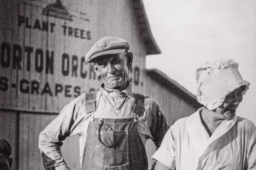 A man in overalls and a cap stands next to a woman in a white dress, with a child in front, against a barn's vintage advertisement.