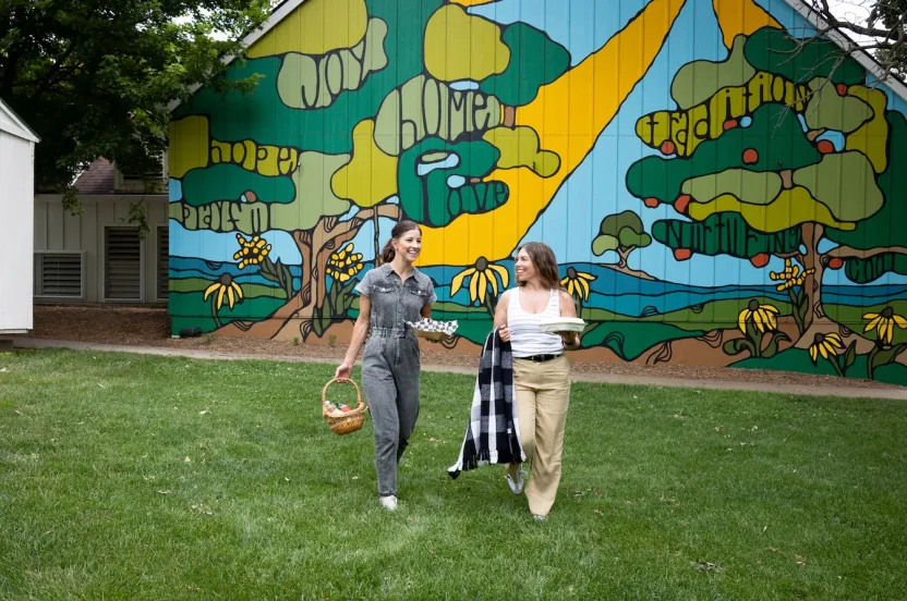 Two women walk on green grass, carrying a basket and a plate, with a colorful mural of trees and landscape behind them.