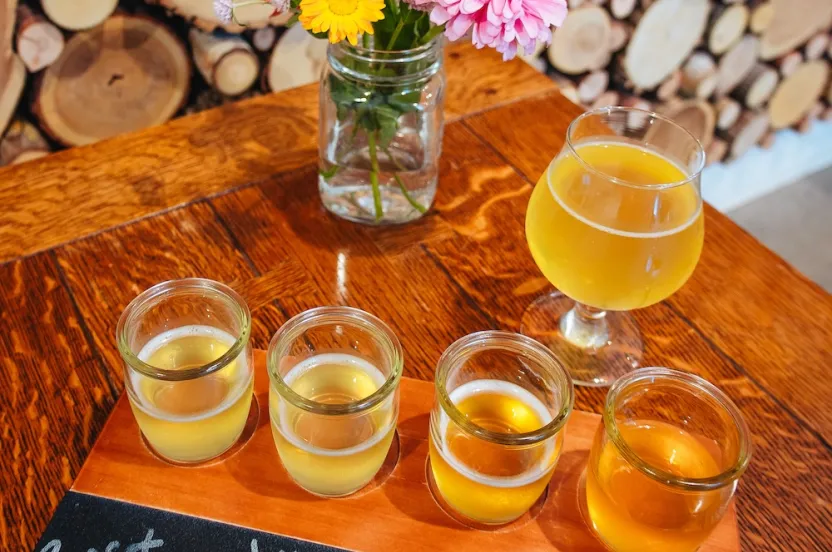A tasting flight of four light drinks labeled 'Russet', 'Joy', 'Tropical Sage', and 'Orange' beside a floral vase on a wooden table.