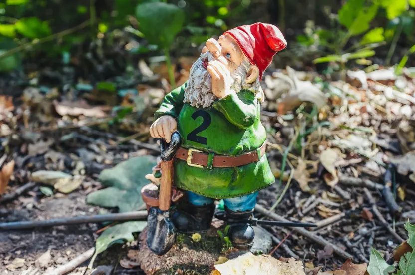 A garden gnome with a red hat and green shirt marked with number 2 stands on soil, holding a shovel. Sunlight filters through surrounding foliage, evoking whimsy.