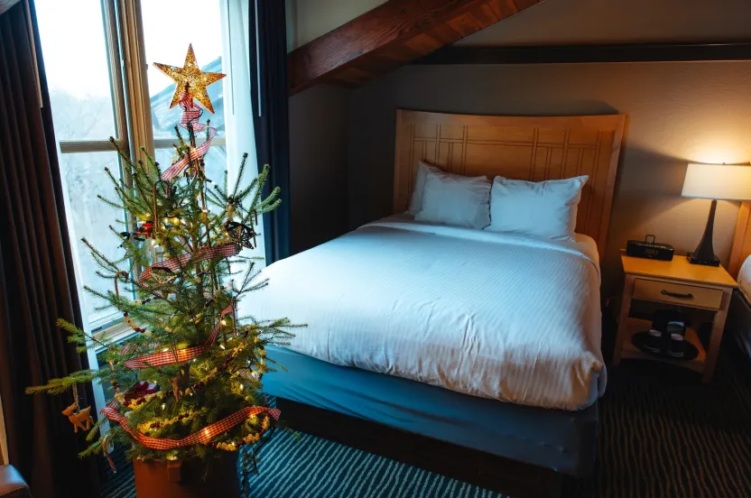 A cozy hotel room featuring a decorated Christmas tree with a star topper, beside a neatly made bed and a bedside lamp.