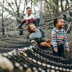 Three children play on a large outdoor net structure, surrounded by trees, engaged in a fun and adventurous activity.