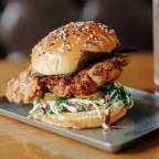 A towering crispy fried chicken sandwich with sesame seed bun, pickles, and slaw, served on a stainless steel plate.