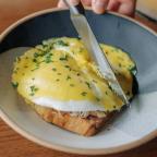 A hand holding a knife cutting into a plate of toasted bread topped with poached eggs and creamy hollandaise sauce, garnished with chives.